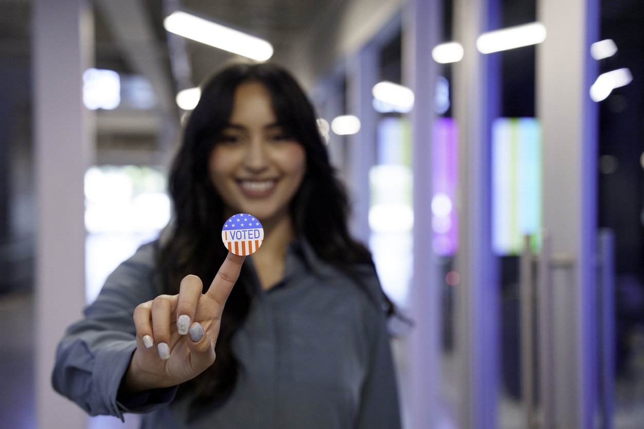Young Hispanic Woman in Her Twenties Proudly Displaying Her I Voted Sticker in a Co-Working Space Signifying That She Has Done Her Civic Duty By Voting For Candidates and Issues of Her Own Choice